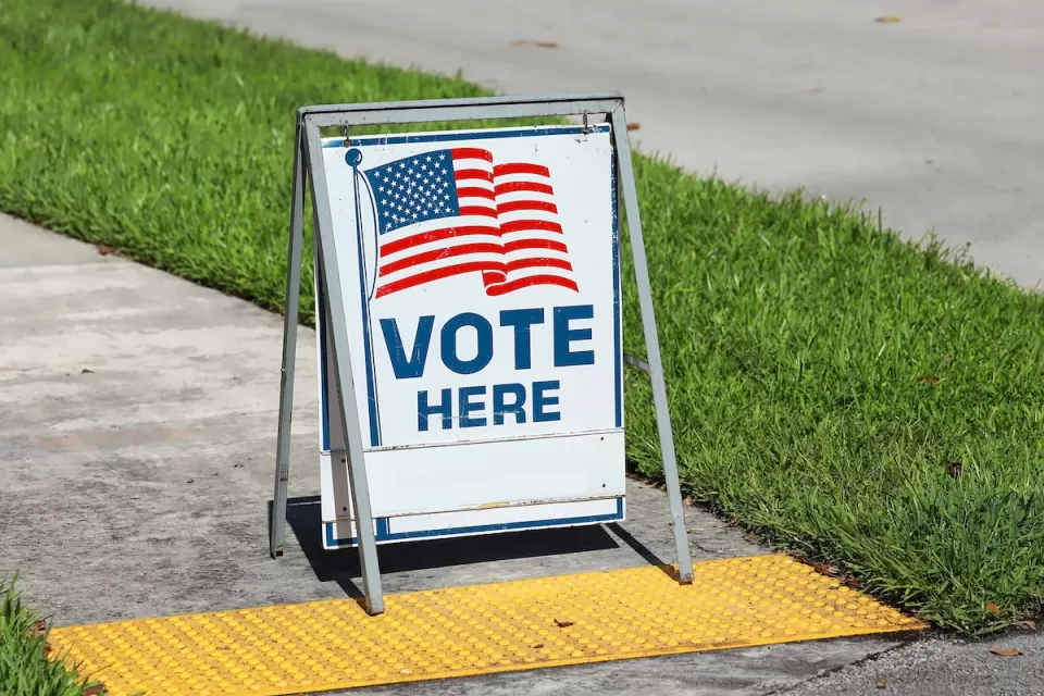 "Vote Here" sandwich board sign with American flag on sidewalk in Fort Lauderdale, Florida.